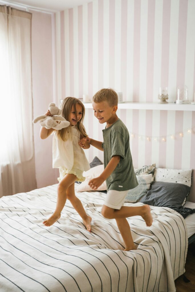 Two siblings happily jumping on a bed in a cozy room, enjoying playful fun and laughter.